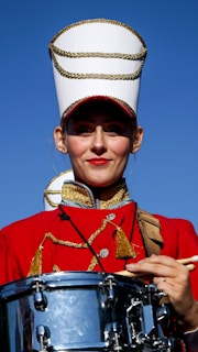 A person is dressed in a red marching band uniform adorned with gold trim. They are wearing a tall white hat with gold details and are holding a snare drum. The background features a clear blue sky.