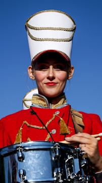 A person is dressed in a red marching band uniform adorned with gold trim. They are wearing a tall white hat with gold details and are holding a snare drum. The background features a clear blue sky.