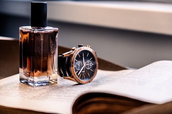 A bottle of perfume and a wristwatch are placed on an open book. The perfume bottle is rectangular with a dark cap, containing a brownish liquid. The wristwatch has a black face and leather strap, with a shiny metallic case and detailed dials.