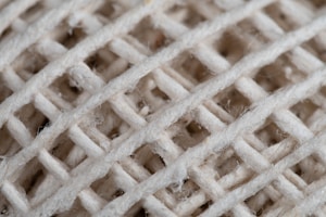 A close-up view of braided or woven material, showing the intricate texture and crisscross pattern of the threads. The fibers appear natural, with some fraying and irregularities visible along the surface.
