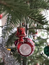 A close-up shot of a Christmas tree adorned with festive decorations, including a red ornament with intricate white patterns and a glass center, surrounded by lush green pine needles. Tinsel and colorful string lights add sparkle and warmth to the scene.