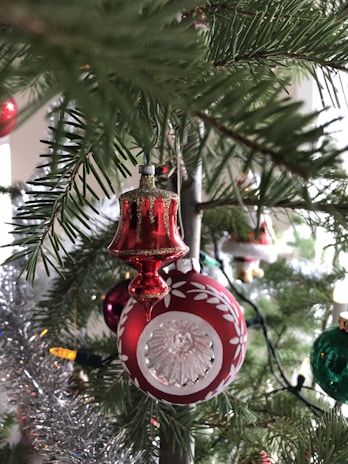 A close-up shot of a Christmas tree adorned with festive decorations, including a red ornament with intricate white patterns and a glass center, surrounded by lush green pine needles. Tinsel and colorful string lights add sparkle and warmth to the scene.