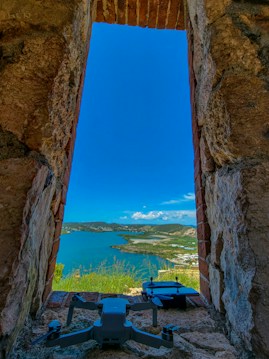 A scenic view of a coastline with a bright blue sky and a vibrant blue sea. The scene is framed by a stone window with rough, textured bricks. Two drones are positioned in the foreground on the stone ledge, adding a modern element to the rustic setting.