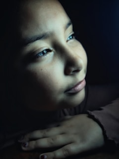 A close-up of a young person's face partially illuminated by soft light, highlighting their thoughtful expression. The person is looking off into the distance with a calm, introspective demeanor. Their hand is resting gently, adding a sense of stillness to the scene.