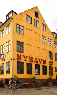 A yellow, multi-story building with large windows and the red lettering 'NYHAVN 17' on the facade. The street in front features people walking and small green plants in wooden barrels beside outdoor seating. The architecture suggests a historic or tourist area.