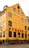 A yellow, multi-story building with large windows and the red lettering 'NYHAVN 17' on the facade. The street in front features people walking and small green plants in wooden barrels beside outdoor seating. The architecture suggests a historic or tourist area.