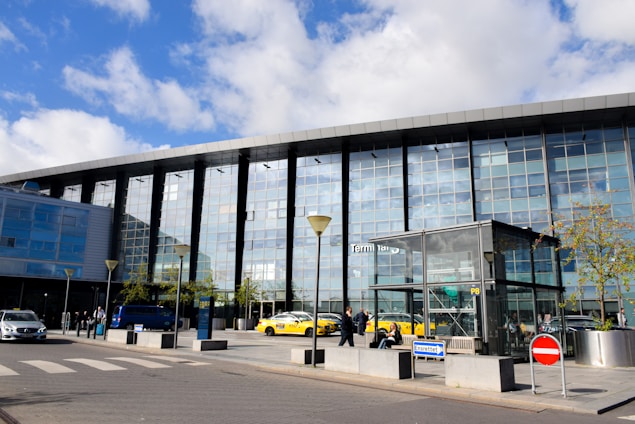 A modern building with a large glass facade reflecting a partly cloudy sky. Yellow taxis are parked in front of the structure, and several people can be seen walking or standing nearby. The area is illuminated by sunlight, and there are some young trees and signs around.