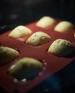 A close-up view of partially baked madeleine cookies in a red silicone baking mold, with a focus on the texture of the batter rising in the oven.