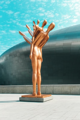 A modern sculpture of elongated human figures, seemingly merging together, stands outdoors against a backdrop of a futuristic building with a smooth, metallic surface. The sculpture is made of a glossy, bronze-like material and is positioned on a minimalist plinth on a paved surface. The sky is bright and partly cloudy.