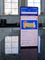 A modern smartphone displaying a shopping app with a focus on winter savings offers, set against a kitchen background. The smartphone screen shows an app interface with search options and promotional content. Sunlight is filtering through large windows, casting reflections on a dark kitchen countertop.