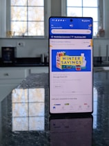 A modern smartphone displaying a shopping app with a focus on winter savings offers, set against a kitchen background. The smartphone screen shows an app interface with search options and promotional content. Sunlight is filtering through large windows, casting reflections on a dark kitchen countertop.