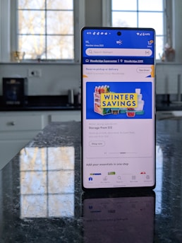 A modern smartphone displaying a shopping app with a focus on winter savings offers, set against a kitchen background. The smartphone screen shows an app interface with search options and promotional content. Sunlight is filtering through large windows, casting reflections on a dark kitchen countertop.