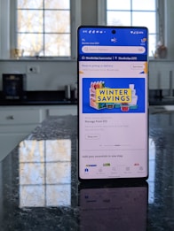 A modern smartphone displaying a shopping app with a focus on winter savings offers, set against a kitchen background. The smartphone screen shows an app interface with search options and promotional content. Sunlight is filtering through large windows, casting reflections on a dark kitchen countertop.