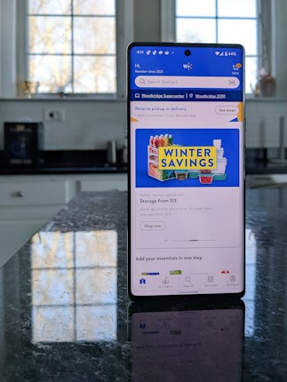 A modern smartphone displaying a shopping app with a focus on winter savings offers, set against a kitchen background. The smartphone screen shows an app interface with search options and promotional content. Sunlight is filtering through large windows, casting reflections on a dark kitchen countertop.