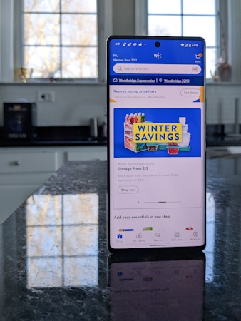 A modern smartphone displaying a shopping app with a focus on winter savings offers, set against a kitchen background. The smartphone screen shows an app interface with search options and promotional content. Sunlight is filtering through large windows, casting reflections on a dark kitchen countertop.