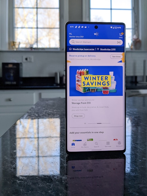 A modern smartphone displaying a shopping app with a focus on winter savings offers, set against a kitchen background. The smartphone screen shows an app interface with search options and promotional content. Sunlight is filtering through large windows, casting reflections on a dark kitchen countertop.