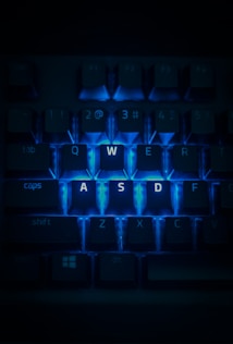 A close-up view of a computer keyboard with the keys W, A, S, and D highlighted by a bright blue backlighting. The surrounding keys are dimly lit, emphasizing the glowing keys in the center. The image has a dark and moody atmosphere.