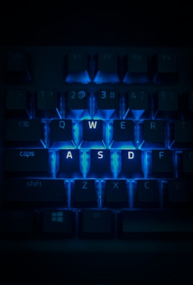 A close-up view of a computer keyboard with the keys W, A, S, and D highlighted by a bright blue backlighting. The surrounding keys are dimly lit, emphasizing the glowing keys in the center. The image has a dark and moody atmosphere.