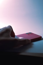 A hand is reaching towards a red smartphone with dual cameras placed on a surface. The phone displays a prominent logo on the back.