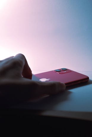 A hand is reaching towards a red smartphone with dual cameras placed on a surface. The phone displays a prominent logo on the back.