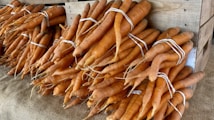 Bundles of fresh carrots are neatly stacked in a wooden crate, with each bundle secured using white rubber bands. The carrots display a rich orange hue and have a rustic, natural appearance. They rest on a surface covered with burlap, emphasizing an organic, farm-fresh setting.