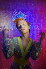 A person with orange hair wearing a sequined outfit with neon accents, posed against a shimmering backdrop. The individual displays unique makeup and an expressive pose, with a headpiece that spells 'FREEDOM'. The lighting creates an ambient, colorful atmosphere with the use of purple and pink tones.