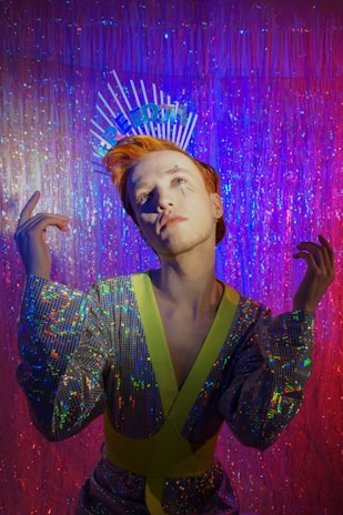 A person with orange hair wearing a sequined outfit with neon accents, posed against a shimmering backdrop. The individual displays unique makeup and an expressive pose, with a headpiece that spells 'FREEDOM'. The lighting creates an ambient, colorful atmosphere with the use of purple and pink tones.