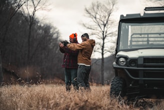 Two people are standing in a field with dry grass and bare trees visible in the background. One of them is wearing an orange beanie and earmuffs, and both are dressed in outdoor clothing. They appear to be holding and aiming a firearm, with one person assisting the other. A utility vehicle is parked nearby, partially visible on the right side of the image.
