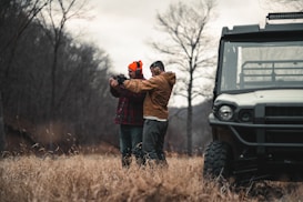 Two people are standing in a field with dry grass and bare trees visible in the background. One of them is wearing an orange beanie and earmuffs, and both are dressed in outdoor clothing. They appear to be holding and aiming a firearm, with one person assisting the other. A utility vehicle is parked nearby, partially visible on the right side of the image.