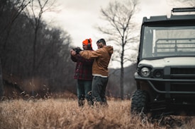 Two people are standing in a field with dry grass and bare trees visible in the background. One of them is wearing an orange beanie and earmuffs, and both are dressed in outdoor clothing. They appear to be holding and aiming a firearm, with one person assisting the other. A utility vehicle is parked nearby, partially visible on the right side of the image.