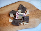 Assorted pieces of chocolate, including dark and peppermint bark, are arranged on a wooden cutting board. The packaging of Ghirardelli Chocolate Squares is visible, surrounded by chocolate with decorative patterns.