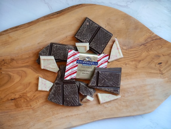 Assorted pieces of chocolate, including dark and peppermint bark, are arranged on a wooden cutting board. The packaging of Ghirardelli Chocolate Squares is visible, surrounded by chocolate with decorative patterns.