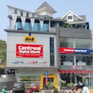 A multi-story modern building with prominent signage, including 'CentreAL Digital World' and various electronic brands such as HP and Samsung. Cars and motorcycles are parked in front, and there's a small garden with plants. The building features large glass windows and a distinctive metal roof design.