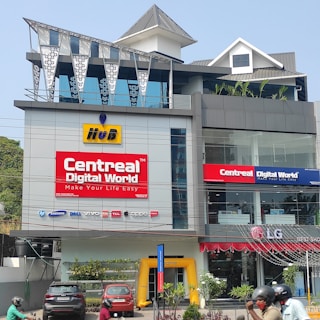 A multi-story modern building with prominent signage, including 'CentreAL Digital World' and various electronic brands such as HP and Samsung. Cars and motorcycles are parked in front, and there's a small garden with plants. The building features large glass windows and a distinctive metal roof design.
