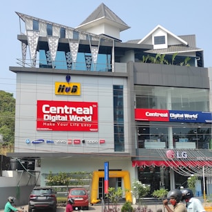 A multi-story modern building with prominent signage, including 'CentreAL Digital World' and various electronic brands such as HP and Samsung. Cars and motorcycles are parked in front, and there's a small garden with plants. The building features large glass windows and a distinctive metal roof design.