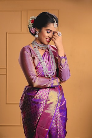 A person dressed in an elegant, traditional purple and pink silk saree accessorized with intricate jewelry, including earrings, necklaces, and rings. The hair is styled with flowers and a decorative headpiece, and the individual appears to be smiling softly while resting one hand near their face.
