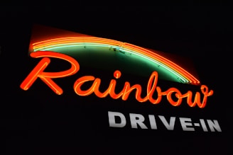A neon sign with the word 'Rainbow' in vibrant red letters. Above the text, a colorful neon rainbow arcs over, featuring shades of red, orange, and green. Below, 'DRIVE-IN' is displayed in bold white letters against a dark background.