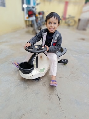 A young child is sitting on a small ride-on toy car. The child is smiling widely and appears very happy. The toy car is black and white with cartoon-like designs, and the child is wearing a jacket and light pants. The setting seems to be an outdoor area with a concrete floor and a blurred background that includes a motorcycle.