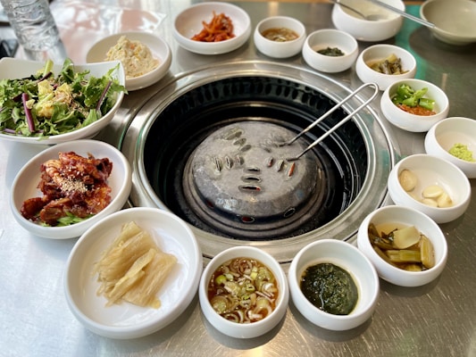 A table setting for a Korean barbecue meal with various small bowls of side dishes including salad, kimchi, pickled vegetables, and sauces arranged around a central grill. The grill has a metal dome and a pair of tongs resting on it.