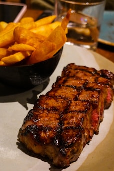 A perfectly grilled steak with distinct char marks is placed on a plate next to a bowl filled with crispy golden French fries. The setting is a cozy dining environment, with reflections of light adding warmth to the scene.