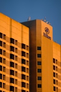 A large hotel building with a modern architectural design is bathed in warm, golden sunlight. The facade features a grid of windows and balconies, and the hotel's logo is prominently displayed at the top corner.