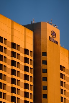 A large hotel building with a modern architectural design is bathed in warm, golden sunlight. The facade features a grid of windows and balconies, and the hotel's logo is prominently displayed at the top corner.