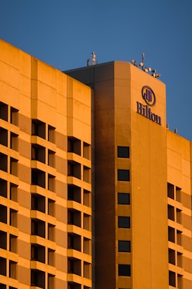 A large hotel building with a modern architectural design is bathed in warm, golden sunlight. The facade features a grid of windows and balconies, and the hotel's logo is prominently displayed at the top corner.
