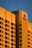 A large hotel building with a modern architectural design is bathed in warm, golden sunlight. The facade features a grid of windows and balconies, and the hotel's logo is prominently displayed at the top corner.
