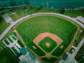 Aerial view of a baseball field surrounded by a road and adjacent to a body of water. The field has a well-defined infield and outfield, with green grass and brown dirt. Light poles are positioned around the perimeter, and nearby there are buildings likely used for facilities or storage.