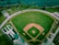 Aerial view of a baseball field surrounded by a road and adjacent to a body of water. The field has a well-defined infield and outfield, with green grass and brown dirt. Light poles are positioned around the perimeter, and nearby there are buildings likely used for facilities or storage.