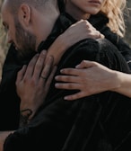 A close-up of two people embracing each other, with one person resting their head against the other's shoulder. Their hands are visible, gently holding each other, conveying a sense of closeness and comfort. The image is softly lit, highlighting their skin tones against the dark fabric of their clothing.
