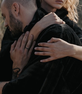 A close-up of two people embracing each other, with one person resting their head against the other's shoulder. Their hands are visible, gently holding each other, conveying a sense of closeness and comfort. The image is softly lit, highlighting their skin tones against the dark fabric of their clothing.