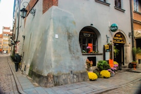 A narrow cobblestone street lined with old buildings features a small pub with a sign advertising music genres like blues and rock. Bright, colorful flowers dot the entrance, and a person walks along the street with their back to the camera. The architecture has an old European charm with arched windows and lantern-style lights.