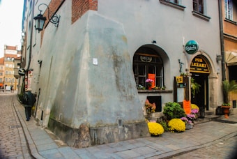 A narrow cobblestone street lined with old buildings features a small pub with a sign advertising music genres like blues and rock. Bright, colorful flowers dot the entrance, and a person walks along the street with their back to the camera. The architecture has an old European charm with arched windows and lantern-style lights.